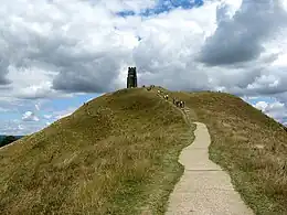 In the background, a square stone church tower atop a steep and narrow terraced hill.  The foreground shows a concrete path leading upwards to the tower. People are hiking up the hill, and in the midground is sheep grazing on the terraces.