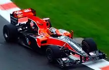 A red and black Formula One car being driven on a damp track near the grass