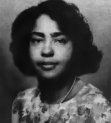 An African-American woman with dark hair side parted, dressed in loose waves to chin length; wearing a floral top or dress