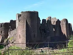 A castle, with a large circular tower facing the viewer, with an angular spur jutting out from the base of the tower; a metal fence is in the foreground, with green vegetation around it.