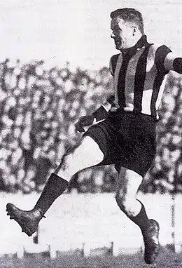 A dark-haired footballer in a kicking motion wearing a long-sleeve black-and-white vertically-striped guernsey, dark shorts and football boots