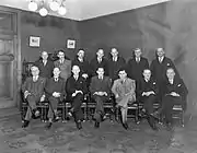 A black-and-white photo of fourteen gentlemen, seven sitting and seven standing behind, all in suits.