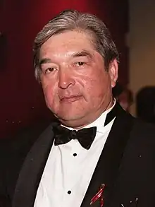 A 45-year-old man with gray hair dressed in a suit smirking towards the camera.