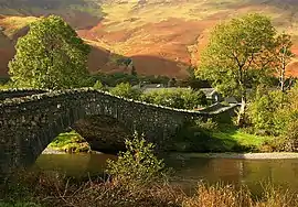 A stone humpback bridge over a small river in front of a green and brown moorland hillside.