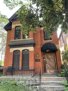 A two-storey brown brick building in the Summertime with a tree covering part of it.