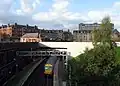 View looking west, to station building to the left of the new road bridge and a train at the eastbound platform. The James Watt College Finnart Campus stands behind houses to the right.