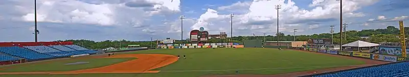 A panorama of the ballpark taken from the right field seats showing the field, entire outfield wall, guitar scoreboard, green trees outside the park, and a partly cloudy blue sky above