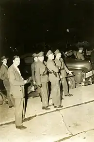 Several policemen with batons stand in front of old vehicles