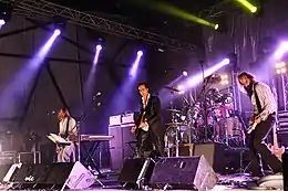 A rock band performing live on-stage. The stage is brightly lit on the left, a man plays bass behind a keyboard. In the centre, a man plays guitar and behind him a man plays drums. On the right, a man plays an electric mandolin.