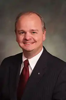 A middle-aged white man is wearing a dark suit, white shirt, and red tie; he has thinning brown hair and is smiling while looking into the camera.