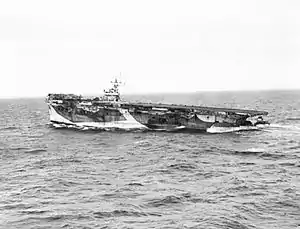 Black and white photograph of an aircraft carrier at sea. The stern of the ship is much lower in the water than the bow.