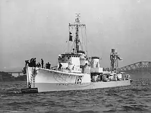 A light coloured warship with two funnels is at anchor. The letters I85 are painted on the ship's bow. A single gun is visible forward. The ship has two masts, with the forward, cruciform, mast carrying a large number of aerials, while the aft lattice mast carries a cylindrical antenna.