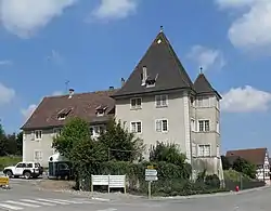 A grey building, in two sections, one two storeys tall with tall, angled, brown tiled roof, the other two intersecting square towers, three storeys tall, each with tall square-pyramidal slate roofs.