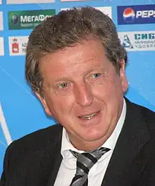 A photograph of a grey-haired, middle-aged man at a press conference. He is wearing a black suit, a white shirt and a grey and black striped tie. He is speaking with someone out of frame.