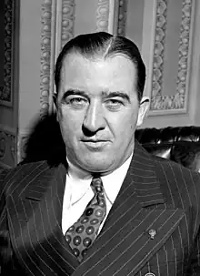 A man with dark, slicked-back hair wearing a pinstriped black jacket, patterned tie, and white shirt