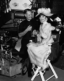 man standing, woman seated, smiling, on tall director's chair in period dress and hat