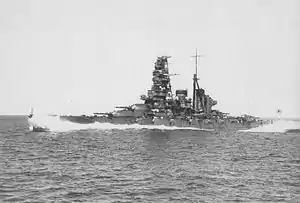 A portside view of Haruna – a large warship with a tall superstructure and two funnels – steaming at full speed in high seas with waves coming over the bow