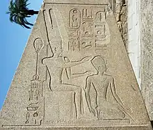 A trapezoidal stone obelisk engraved with Egyptian hieroglyphs and two figures. The figure on the left is seated and wears a tall crown, both hands outstretched to the figure on the right, kneeling, who wears a much smaller crown.