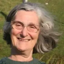 The photograph is a close-up portrait of Helen Steven. Helen is wearing glasses and smiling into the camera. You can't see much of the background as the photo is a close up of her face, but she appears to be in a sunny, green field.