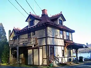 A light yellow building with dark red roof and trim and a brick chimney at the center seen from one of its corners. At bottom right is an overhanging roof with a sign on it saying "Highland Falls". In the rear is a large body of water.