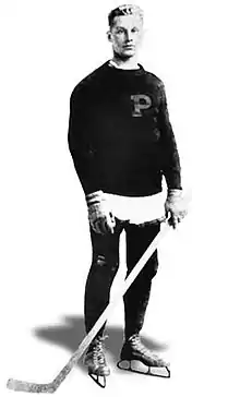 Young man posing for a photo wearing skates and holding a hockey stick. He is also wearing a sweater with a large letter "P" on it