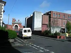 Photo of a large brick building with gate at front. A white van is parked on a road leading to the gate.