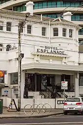 External shot of a two-storey building. The entrance is at left with the name displayed above. The building has a white picket fence in front. Two workmen are visible descending the entrance steps with a section of the same white fencing. To the right of the entrance is a hand-written notice in green-blue writing: "Patrons must be seated at all times". The building's façade is off-white.