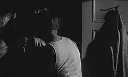 Man in sleeveless undershirt presses a woman against a wall in a dark room