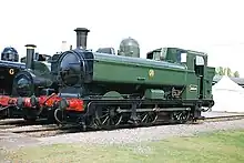 A green pannier tank locomotive, viewed from the left and front, is standing in front of two other locomotives. The locomotive is fitted with a chimney, rounded boiler dome, and a curved safety valve cover. The cab is enclosed with rectangular windows. Although mainly green, the chimney, front, and undercarriage are painted black, except the coupling rods which are grey, and the front buffer beam is painted red. On the side of the pannier tank is a yellow roundel with the letters G W R. On the side of the cab there is a small blue disc and a black number plate with the letters and edging in yellow.