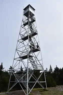 The Fire Tower on top of Hunter Mountain. Stairs lead from the ground going up, oscillating direction as they ascend. The top cabin of the tower is locked. Pine trees in the background sit beneath a slightly overcast sky.