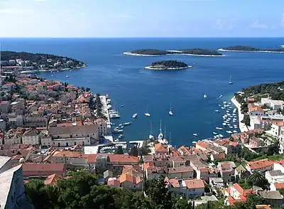 A photograph of a coastline with a light blue sky and white clouds above, a dark blue ocean in the background, and land spotted with pink-roofed, white-walled buildings in the foreground