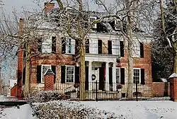 Red brick building with snow on the dark roof. In front of the building, there are trees and snow on the ground.