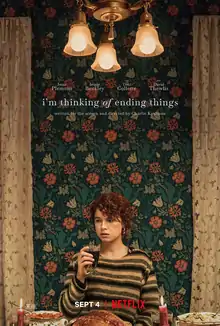 "Promotional release poster": A young woman sits at a dinner table holding a drink. The background features a wallpaper decorated with flowers. Near the top of the poster are three ceiling lamps and the title, "i'm thinking of ending things".