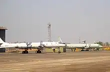 Two four-engine propeller-driven aircraft on ramp facing right of screen. The large aircraft in the midground, which is slightly off-centered, has swept-back wings and is painted in a pale grey paint scheme. The other in the background is a straight-wing aircraft painted in pale green