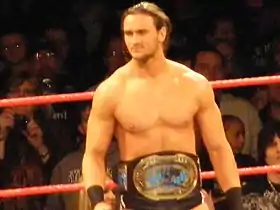 A Caucasian male stands in a wrestling ring with red ropes. He has long dark hair tied back, and is wearing short, black wrestling tights. A championship belt is around his waist, with a black strap and gold center plate.