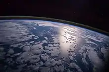 Image 39A view of Earth with different layers of its atmosphere visible: the troposphere with its clouds casting shadows, a band of stratospheric blue sky at the horizon, and a line of green airglow of the lower thermosphere around an altitude of 100 km, at the edge of space (from Earth)