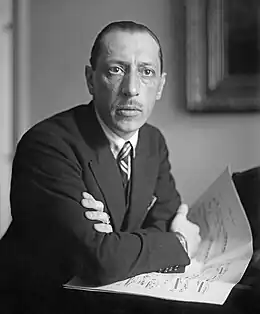 A man with short hair and glasses, wearing a dark suit and leaning against a piano