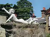 Colour photograph of an individual female statue with short tousled hair lying on a rock pedestal. Supporting herself on her left arm, her head is up and her right arm raised