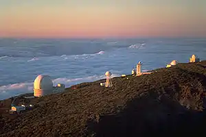 Series of white structures along the side of a mountain with a sea of clouds below and behind the mountain extending to the horizon which is red, orange, and yellow.