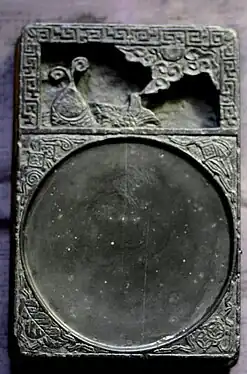 A white, rectangular stone tablet with two sections. The top quarter section contains a carving of a fish, and the bottom three-fourths contain a shallow, circular indent for the ink.