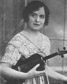 A young white woman with short dark hair, wearing a lace dress and holding a violin.