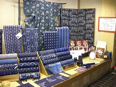 A display table showing a number of different indigo-dyed kasuri fabrics, some stacked on wooden stands in roll-form, others displayed flat in front of the rolls, and some hanging on small stands.