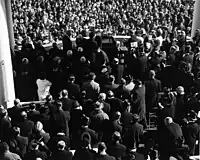 The inauguration of John F. Kennedy as seen from behind. Most men have their hats off; however a few top hats can be distinguished, some by the shininess of the hat's flat crown