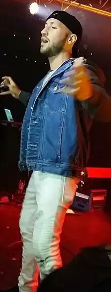 Colour photo of Jonathan Perry on stage wearing a black baseball cap, a blue denim jacket, and white trousers