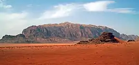 A wide rocky tan-colored ridge, reaching a high point in the middle, over a desert of reddish sand with a smaller prominence in the foreground