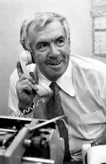 Black and white photo of Beauchamp sitting at a desk, talking on a telephone and using a typewriter