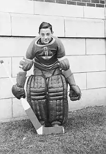 A teenage Plante assumes the traditional goaltender stance, slightly crouched with legs together, wearing goaltender pads on his legs, his team sweater, and holding a goaltender stick in his right hand with the blade of the stick in front of his feet