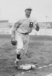 A man in a light baseball uniform with a dark "B" on the left chest running past first base with a fielding glove on his right hand.