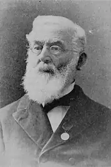 A black-and-white photographic portrait of a bearded, white-haired bespectacled man, shown from the chest up