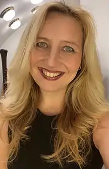 white woman with long blonde hair, wearing a short-sleeved black top, grinning at camera